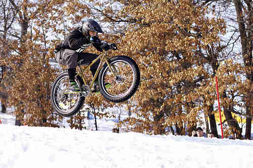 Right side view of a cyclist riding a Surly Pugsley bike, flying through the air, off of a snow mound on a ski hill
